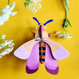 Cardboard sculpture of a bee with pink wings mounted on a bright yellow wall surrounded by Queen Anne's Lace flowers. 