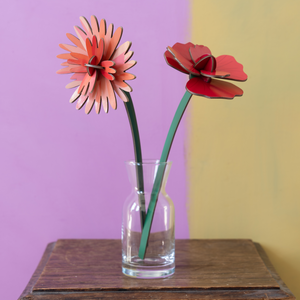 Two cardboard flowers in a small glass vase on a wooden table against a two tone wall. 