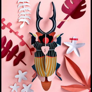 Cardboard sculpture of a giant stag beetle on a ink wall with various pink and white leaves. 