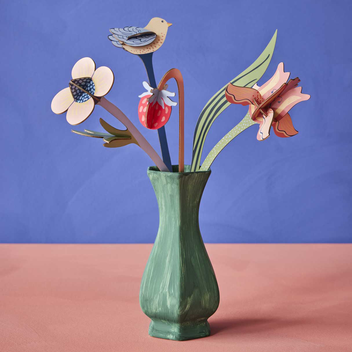 Bouquet of cardboard flowers, a strawberry, and bird in a green painted vase on a pink foreground against a periwinkle background. 