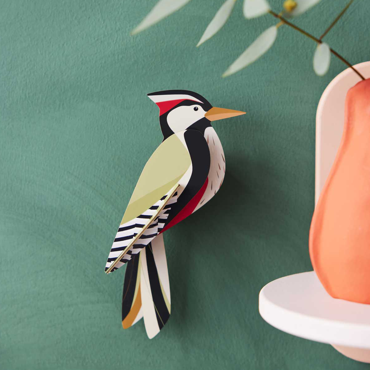 Cardboard sculpture of a woodpecker mounted on a green wall with an orange vase in the foreground.