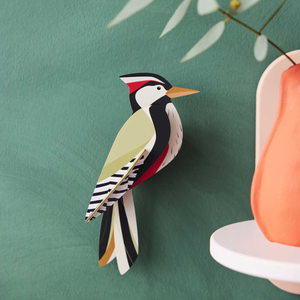 Cardboard sculpture of a woodpecker mounted on a green wall with an orange vase in the foreground.