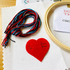 Embroidery hoop, red felt heart with needles in it, and colorful thread on a wooden surface