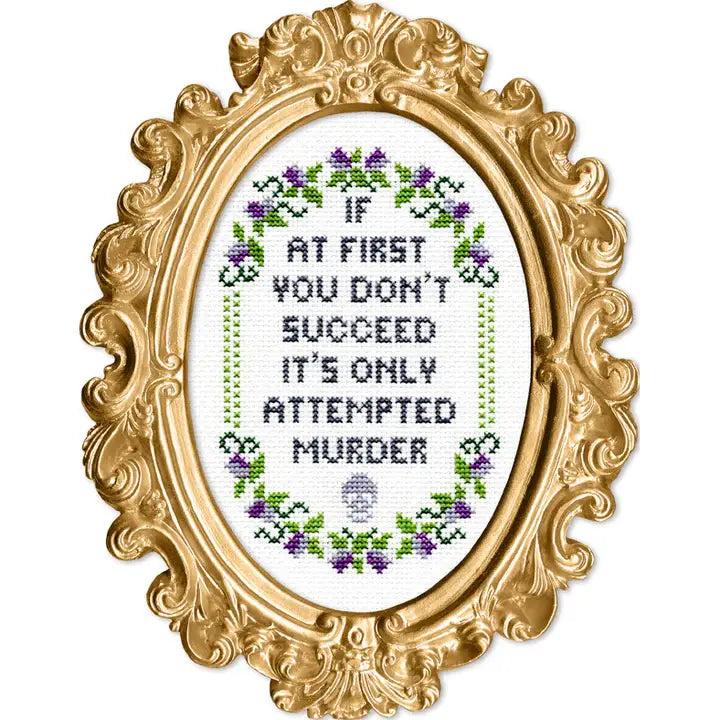 Decorative gold frame with cross-stitched text that says "If at first you don't succeed it's only attempted murder" and floral patterns on a white background