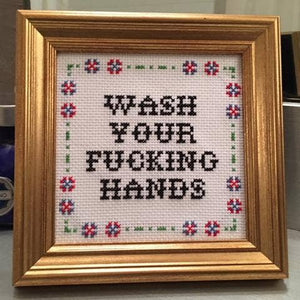 Framed cross-stitched art with text that says "wash your fucking hands"