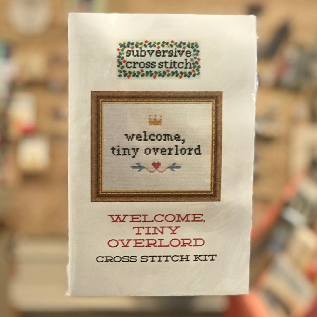 A cross stitch kit from Subversive Cross Stitch in its packaging with a picture of the completed cross stitch, which says "Welcome, tiny overlord" on it with a yellow crown and a red heart with blue leaf shapes. 