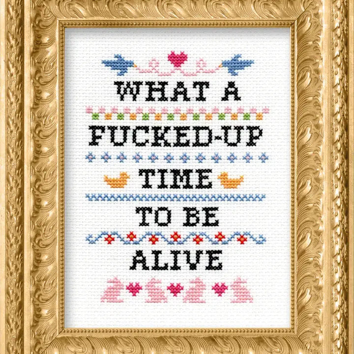Cross-stitched text that says "What a fucked up time to be alive" in a decorative gold frame