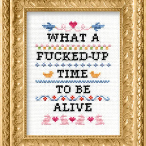 Cross-stitched text that says "What a fucked up time to be alive" in a decorative gold frame