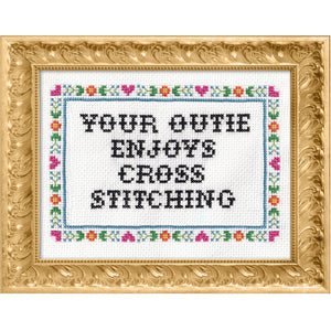 Cross-stitched text that says "your outie enjoys cross stitching" in a decorative gold frame with floral border