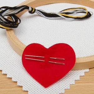 Red heart-shaped fabric piece with needles on a white embroidery hoop.
