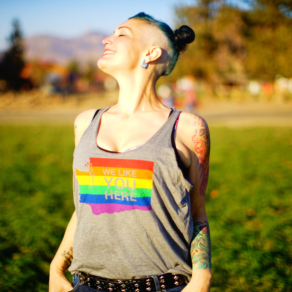 A person smiles up into the sunshine while wearing a light gray tank top that says "We like you here" and has a rainbow in the shape of Washington state.
