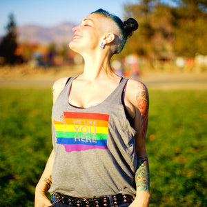 A person smiles up into the sunshine while wearing a light gray tank top that says "We like you here" and has a rainbow in the shape of Washington state.