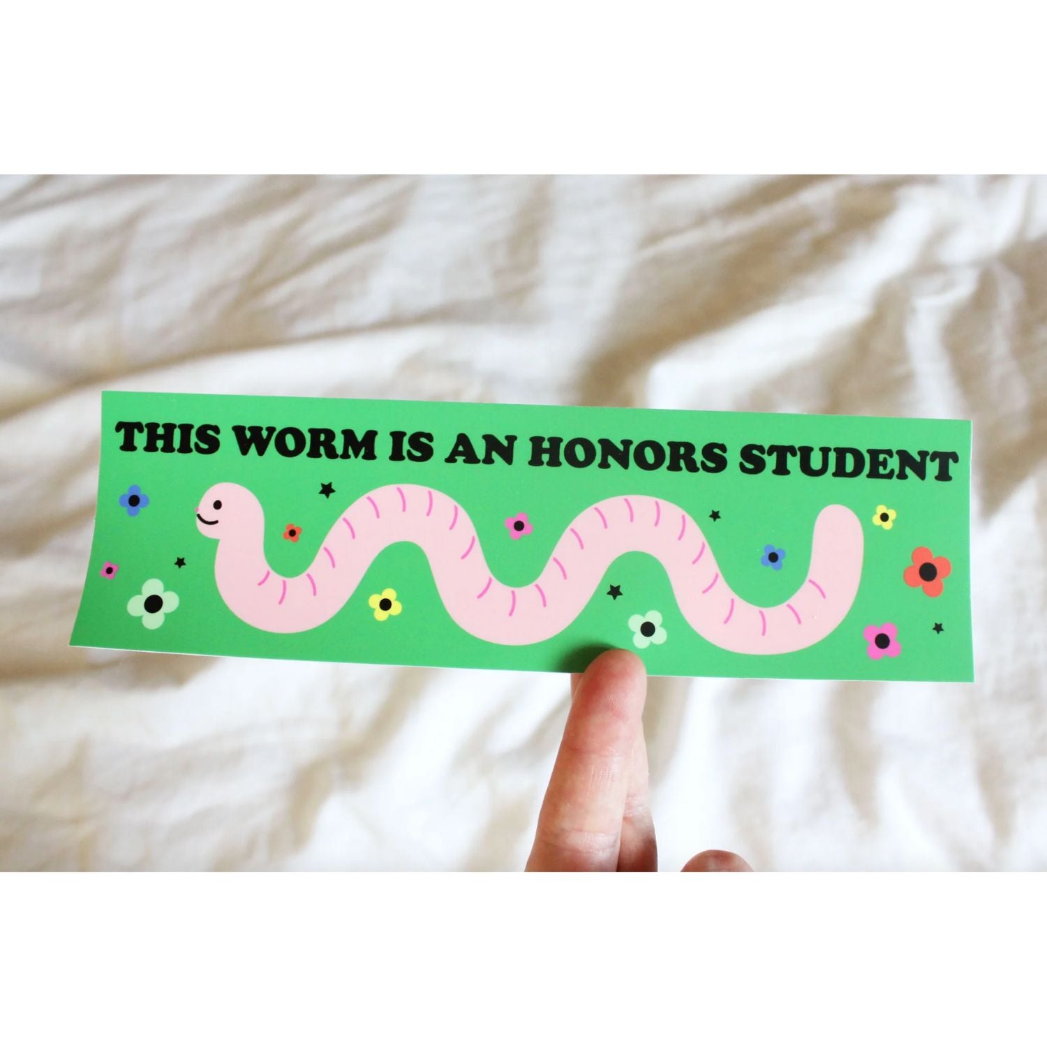 Sticker with a pink worm illustration and text that says "this worm is an honors student" on a green background, held against a white fabric background.