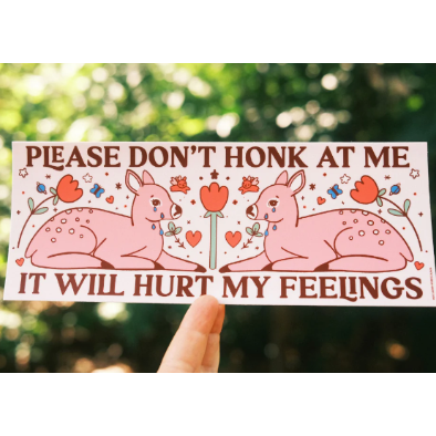Cartoon deer with flowers and text 'Please don't honk at me it will hurt my feelings' held against a blurred green background.
