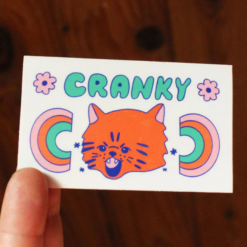 A sticker that says "cranky" in green bubble letters and has an orange cat showing its teeth with two rainbows next to it