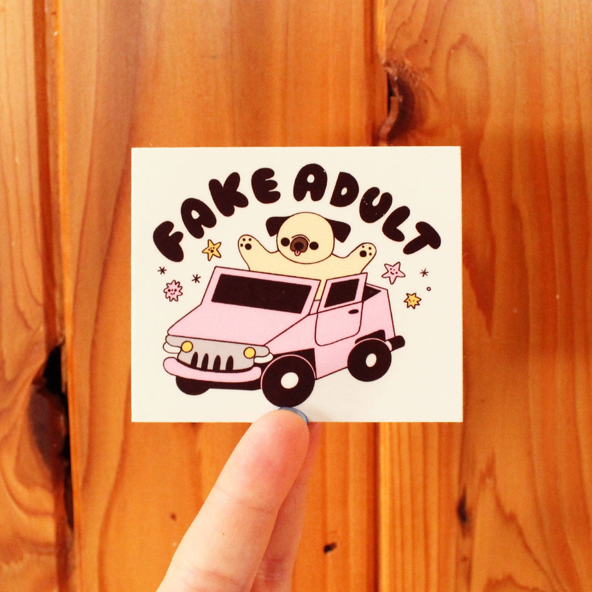 Sticker with a cartoon dog in a car and 'FAKE ADULT' text held against a wooden background