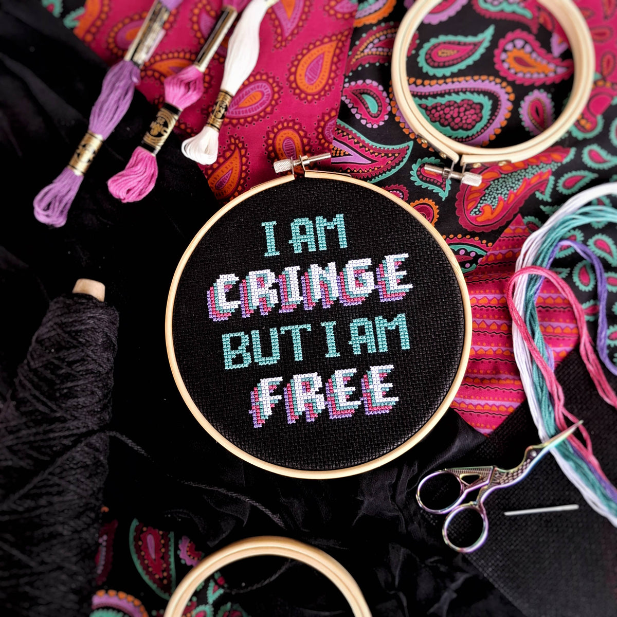 Embroidery hoop with 'I am cringe but I am free' text surrounded by embroidery threads and scissors on a colorful paisley patterned fabric.