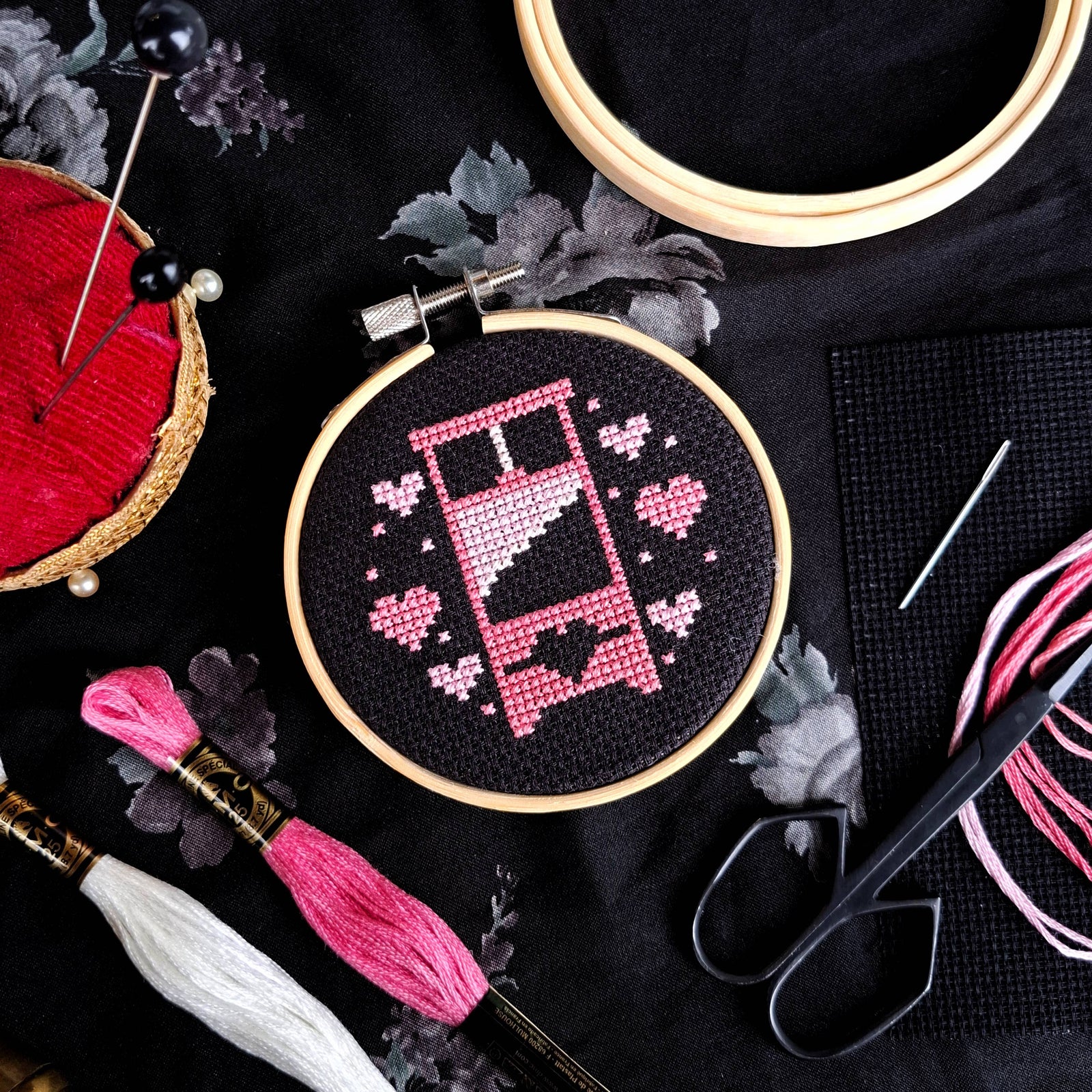Embroidery hoop with pink and white guillotine design on black fabric, surrounded by embroidery tools.