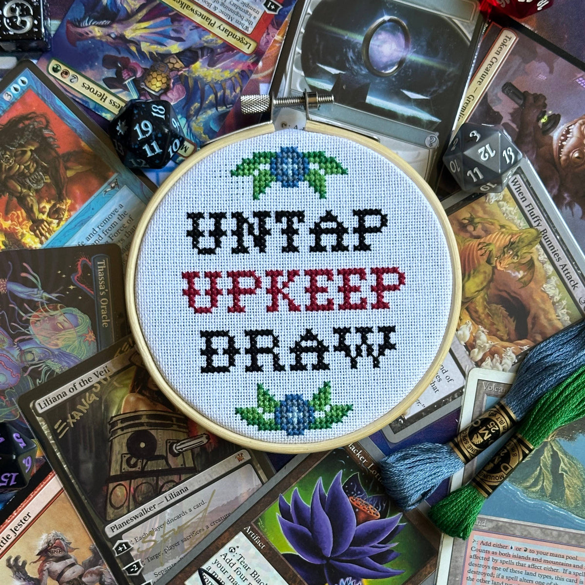 A round cross-stitch embroidery hoop in the center of a tabletop flat-lay, featuring the text 'UNTAP UPKEEP DRAW' stitched in black and red thread with green and blue floral motifs. The hoop is surrounded by fantasy trading cards, dice, and threads on a blue surface.