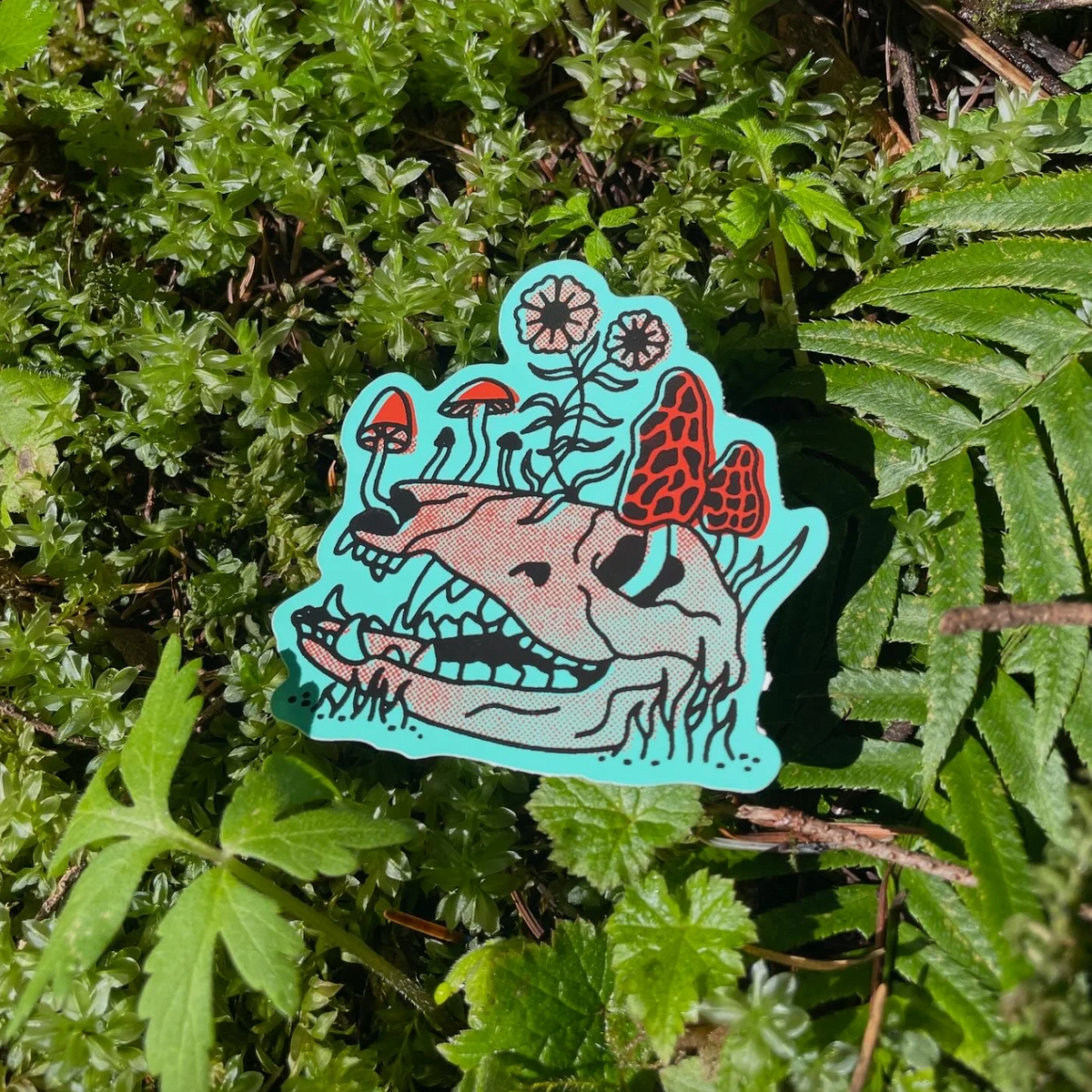 Sticker with a blue background of an animal skull with red mushrooms and flowers growing out of it against a background of fern leaves. 