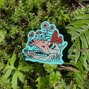 Sticker with a blue background of an animal skull with red mushrooms and flowers growing out of it against a background of fern leaves. 
