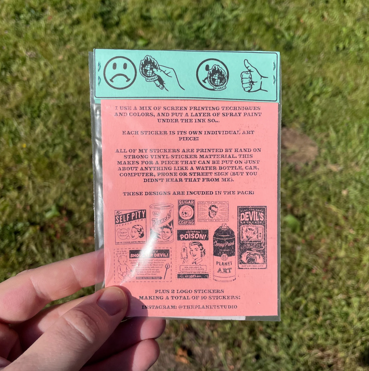 A hand holds a pink and mint green sticker pack against a blurred green grass background. The sticker sheet contains text and several small graphic stickers.