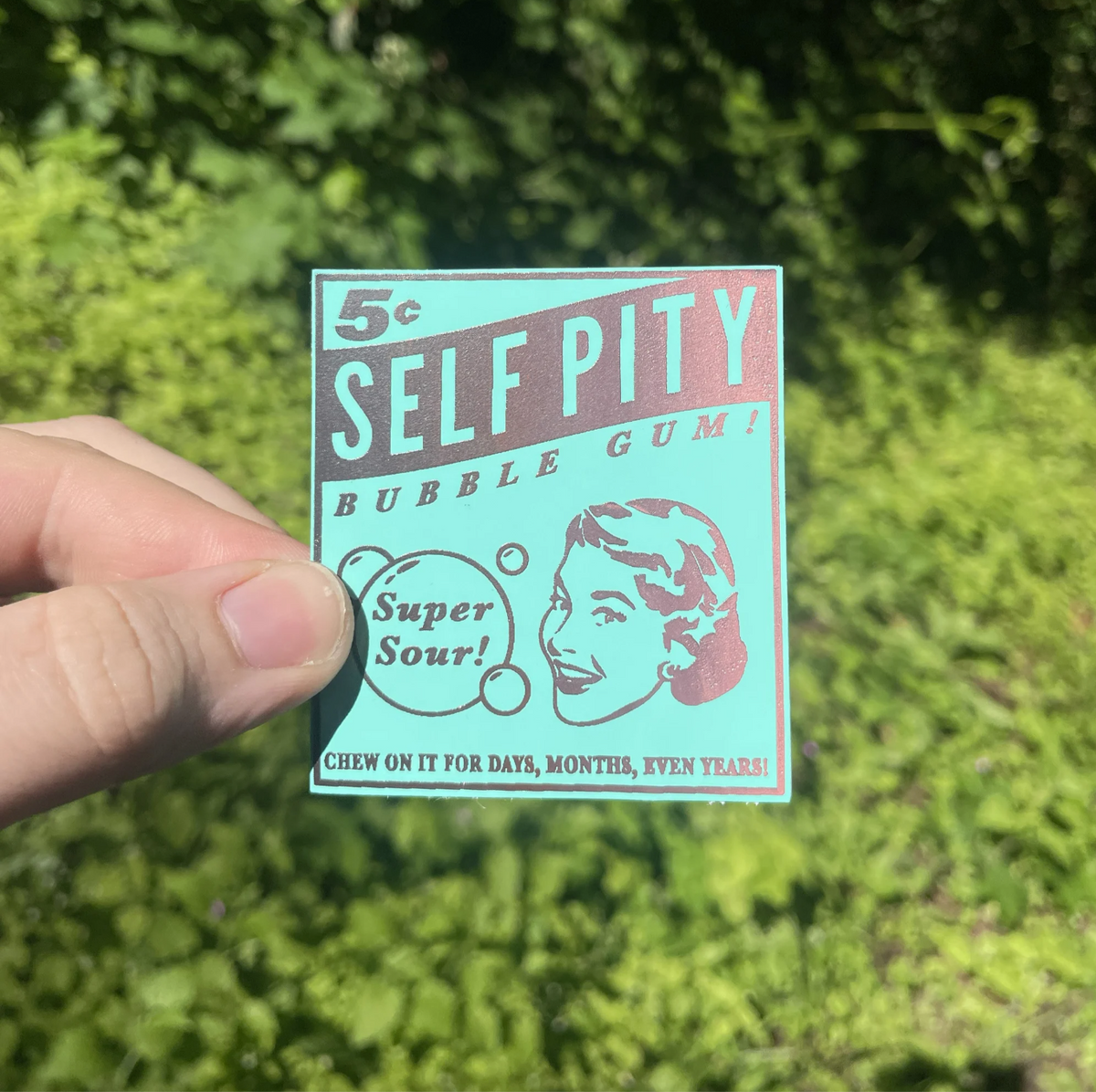 A hand holds a vintage-style square sticker reading '5c SELF PITY BUBBLE GUM!' with 'Super Sour!' and a retro woman illustration, against a blurred green outdoor background.