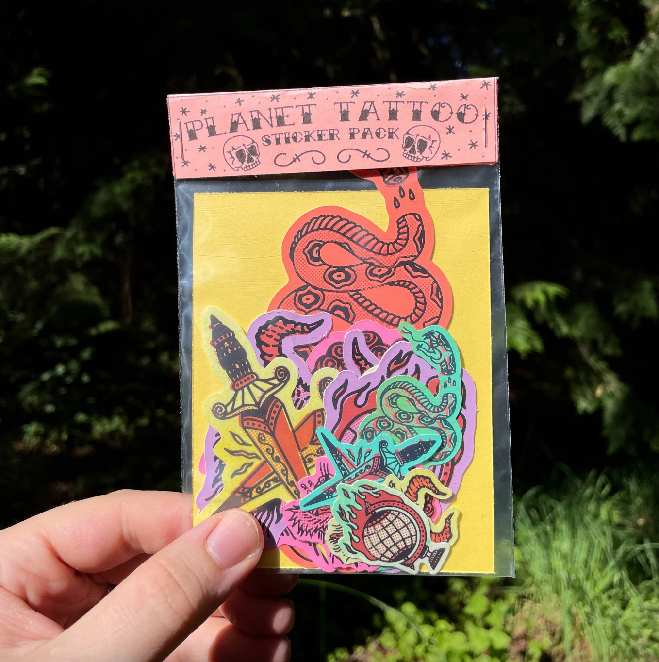 A hand holds a clear plastic package of Planet Tattoo sticker pack against a blurred green outdoor background. The package contains colorful stickers featuring  a snake, dagger, and other tribal-style designs on a yellow backing card.