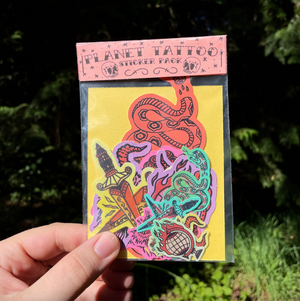 A hand holds a clear plastic package of Planet Tattoo sticker pack against a blurred green outdoor background. The package contains colorful stickers featuring  a snake, dagger, and other tribal-style designs on a yellow backing card.
