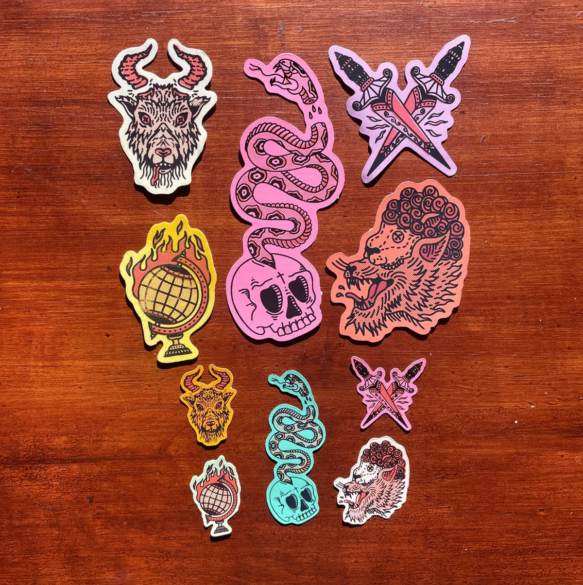 A set of colorful die-cut stickers featuring gothic and edgy illustrations, arranged on a dark brown wooden surface. The designs include a horned goat head, a pink snake with a skull and eye motif, crossed knives with a lightning bolt, a yellow globe with flames, a pink skull, and a lion head.