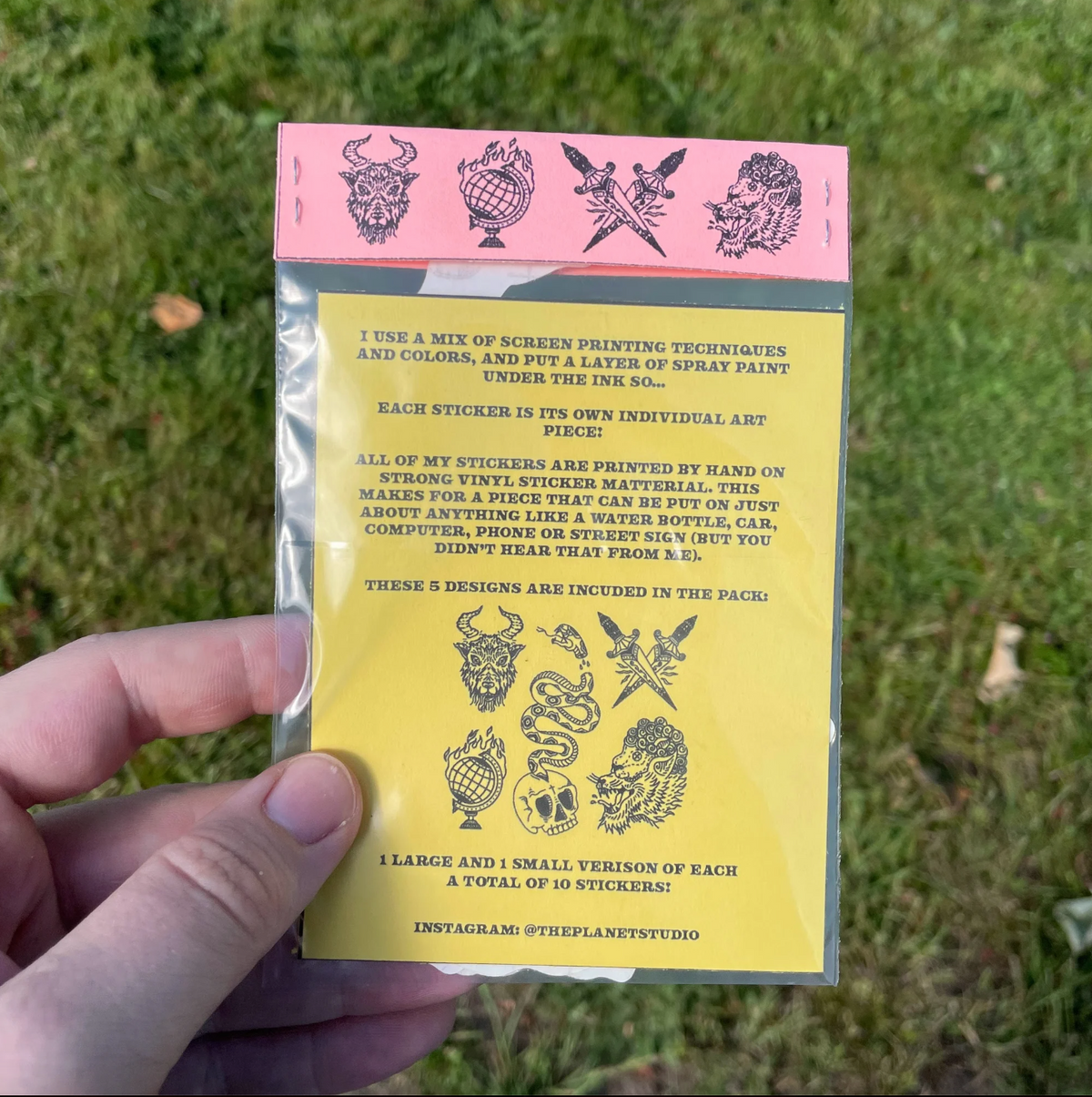 A hand holds a yellow and pink sticker pack card outdoors on grass. The card shows black illustrated designs and printed information about the sticker set, with a visible Instagram handle.