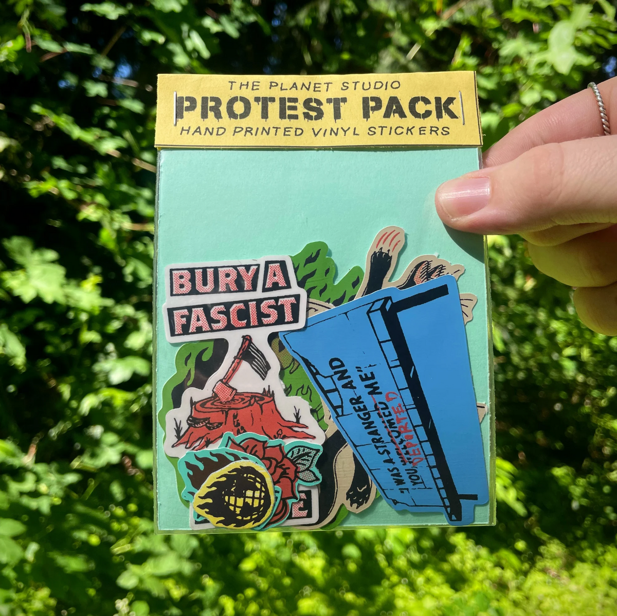 A hand holds a package of 'The Planet Studio Protest Pack' vinyl stickers against a background of green outdoor foliage. The package contains multiple colorful stickers with political and social messages.