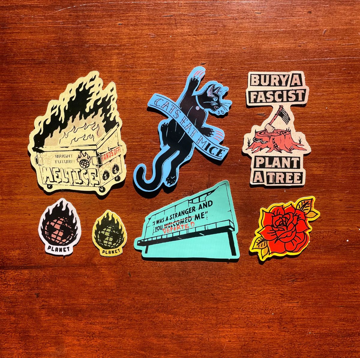 A set of seven colorful political and social stickers arranged on a brown wooden surface. The stickers feature various designs including a black cat with a banner, a burning dumpster, and a red rose. 
