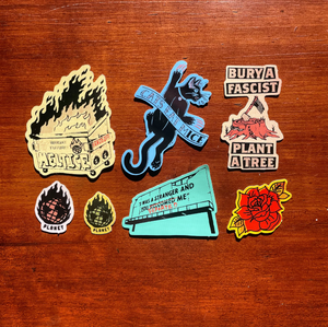 A set of seven colorful political and social stickers arranged on a brown wooden surface. The stickers feature various designs including a black cat with a banner, a burning dumpster, and a red rose. 