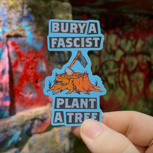 A hand holding a die-cut sticker with the text 'BURY A FASCIST' at the top and 'PLANT A TREE' at the bottom, featuring an illustration of an axe embedded in a tree stump. The background is a blurred graffiti-covered wall.
