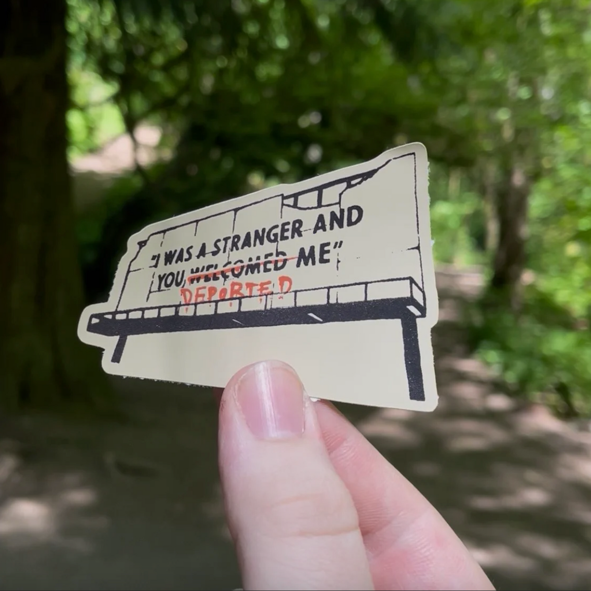 A hand holds a die-cut sticker featuring a stylized billboard with the text “I WAS A STRANGER AND YOU WELCOMED (struck through) DEPORTED ME” in black and red lettering, set against a blurred outdoor forest background.