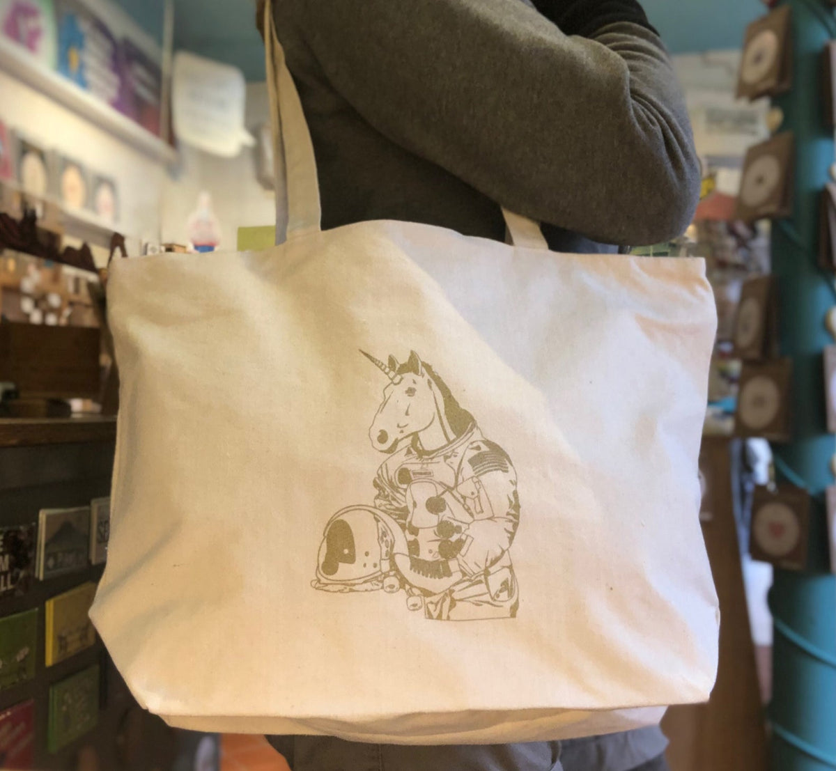 A beige cotton canvas tote bag with a screen print of an astronaut unicorn character, featuring a zipper closure and a 30-inch handle.