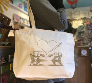 A person modeling a cream colored zippered tote bag with the Love AT-AT First Sight design printed on it in gold ink. 
