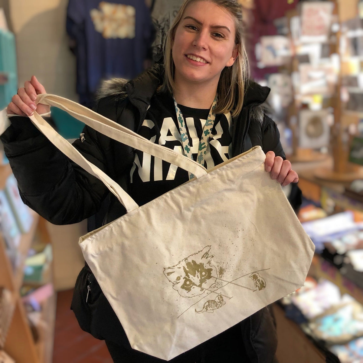 A smiling person holding up a natural color tote bag with a cat design printed in gold ink. 