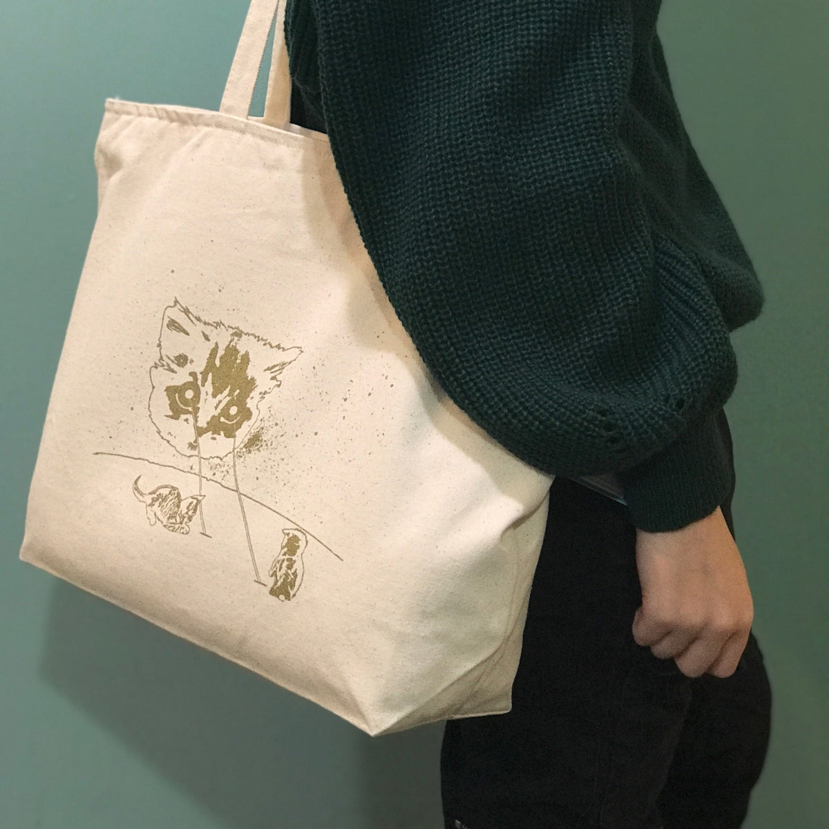 A person carrying a beige cotton tote bag with a gold screen printed cat design on one side.