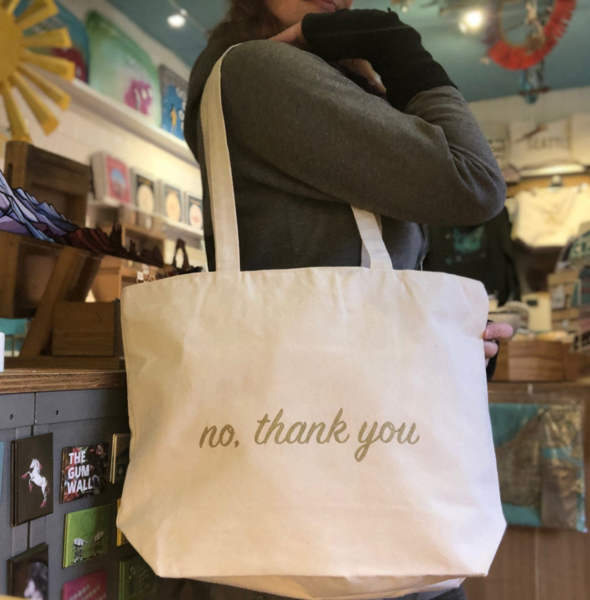 A person modeling a cream colored tote bag that says "no, thank you" in gold ink 