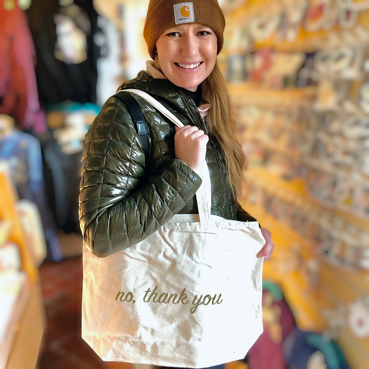 A smiling person holding a cream colored tote bag that says "No, thank you" in gold ink. 