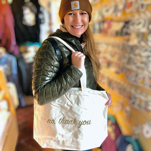 A smiling person holding a cream colored tote bag that says "No, thank you" in gold ink. 