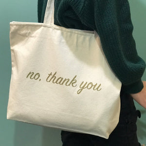 A cream colored tote bag with "No, thank you" printed in gold ink. 