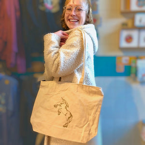 Smiling person holding a cream colored tote bag with a roller skating unicorn printed in gold ink. 