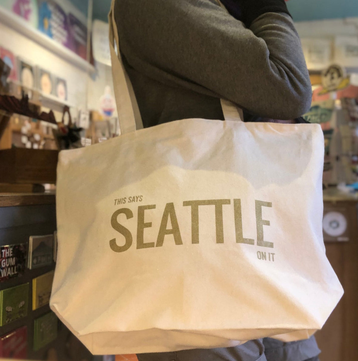 A person holding a cream colored tote bag that says "This says Seattle on it" in gold ink. 