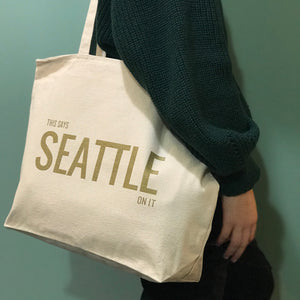 A person carrying a white cotton tote bag with 'This Says Seattle On It' printed in gold on the front side, with a zipper closure.