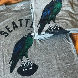 A stack of folded t-shirts that say Seattle and have a hawk carrying a football depicted on them. 