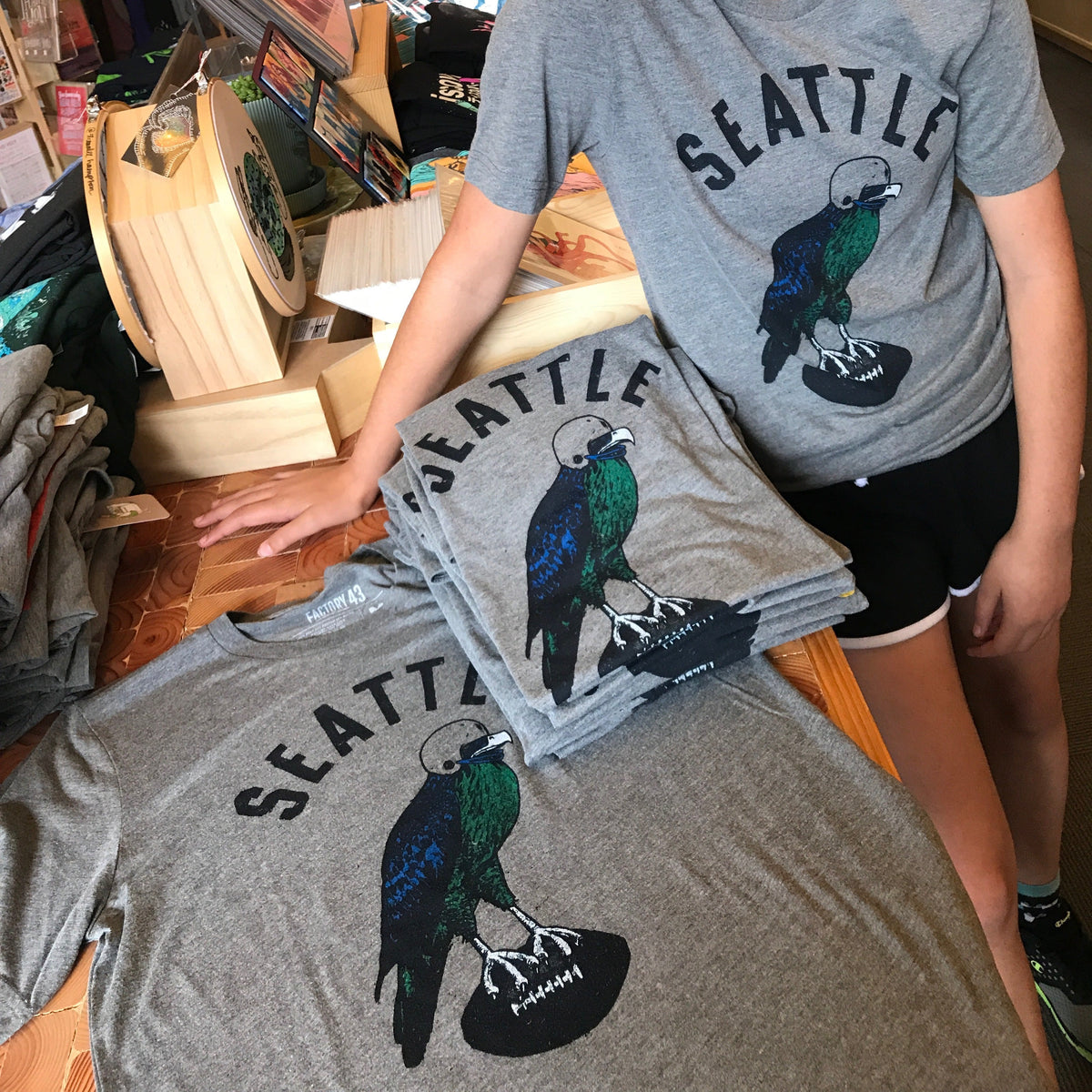 A stack of folded t-shirts that say Seattle and have a hawk carrying a football depicted on them. A young person is modeling the xs size shirt.