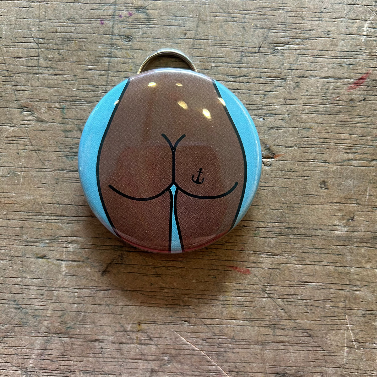Bottle opener keychain featuring a design of a brown butt with an anchor tattoo on a blue background on top of a wooden surface. 